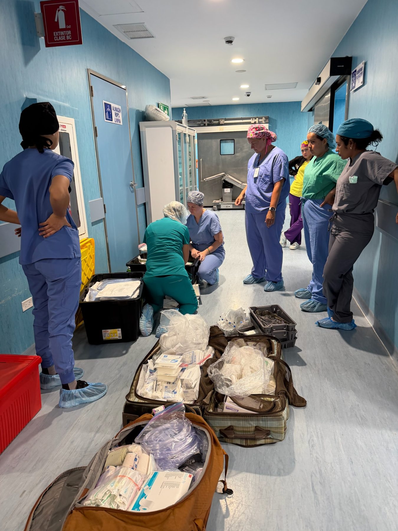 Team sorting surgical instruments and supplies from suitcases in a hospital hallway marked ALMACEN ROPA LIMPIA