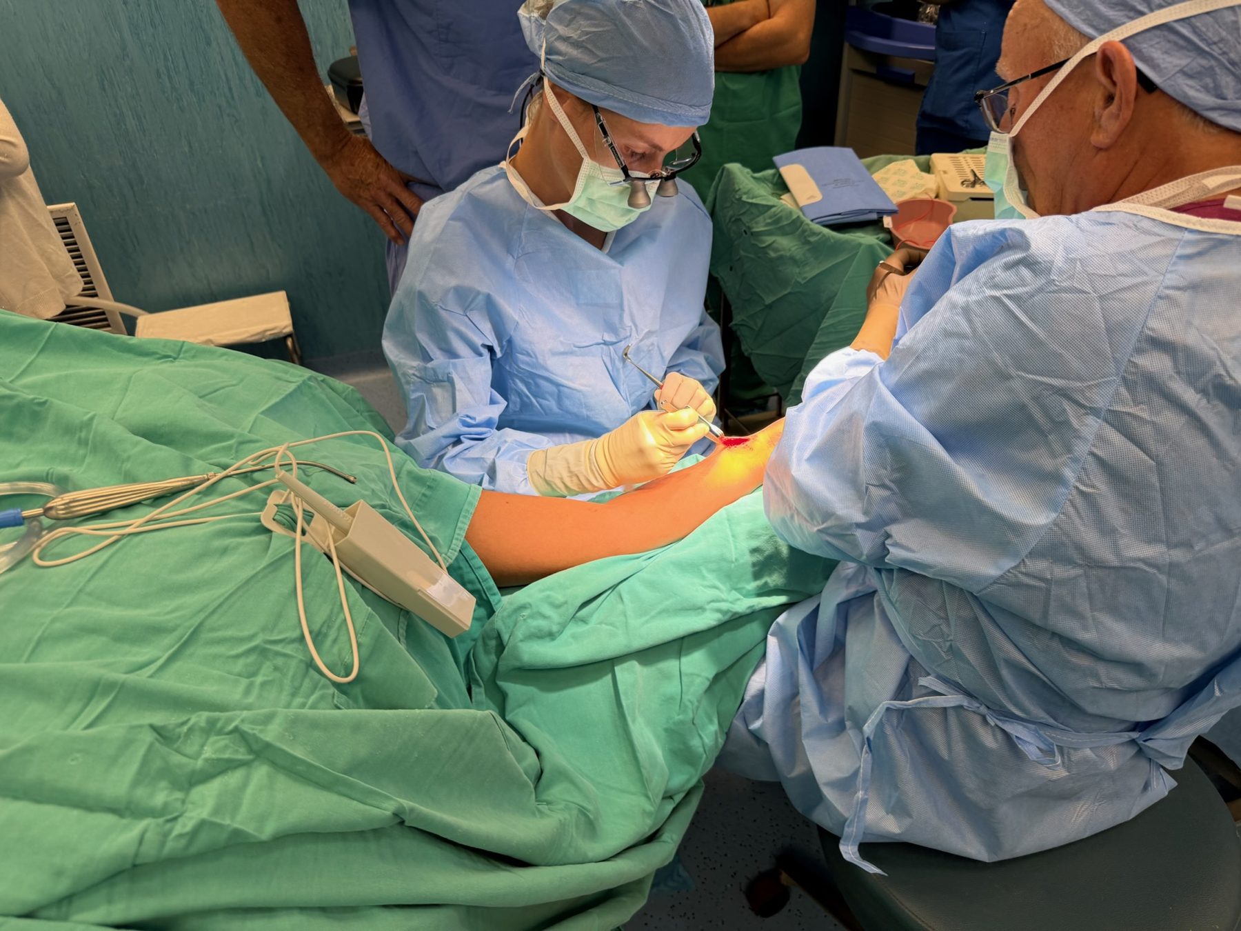 Two surgeons wearing loupes operating on an arm; patient face not shown