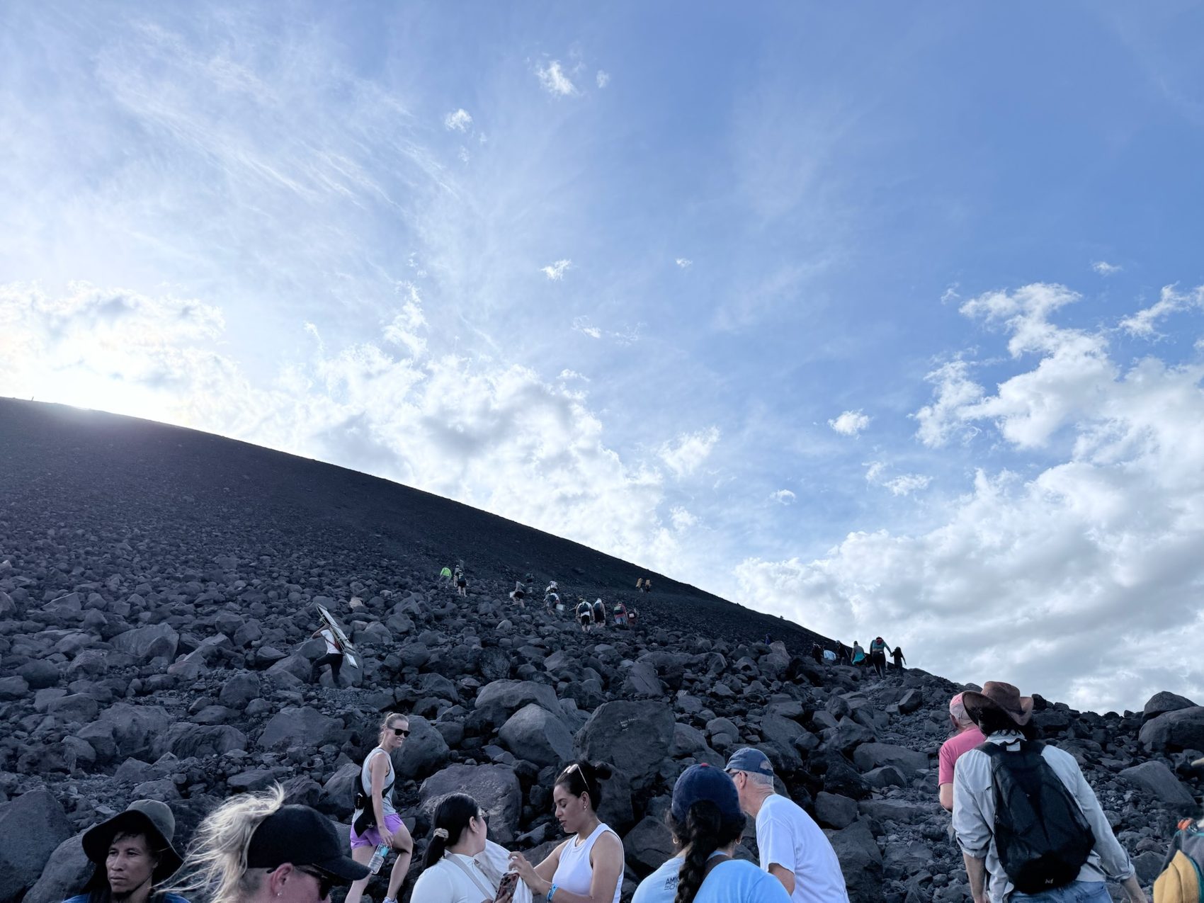 Looking up a black volcanic ridge with hikers and a board against a bright sky