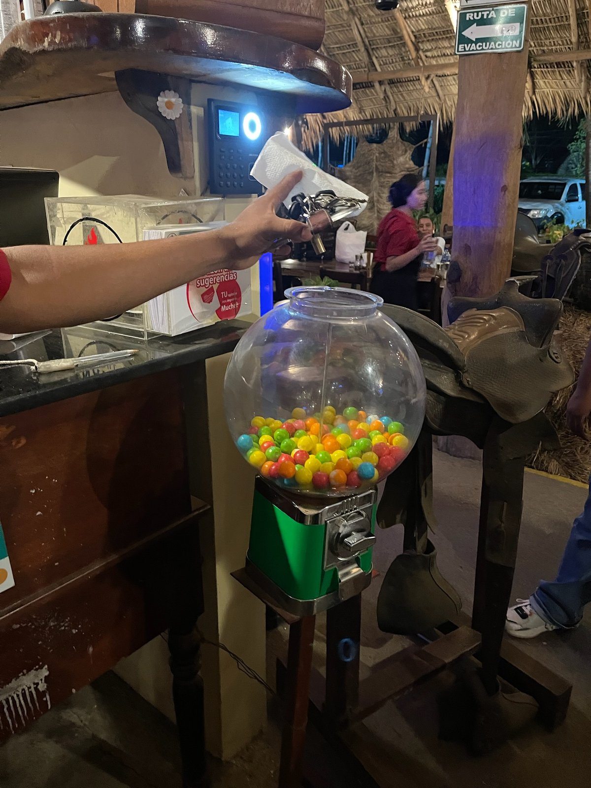 Hand unlocking a green gumball machine under a thatched roof with a RUTA DE EVACUACIÓN sign