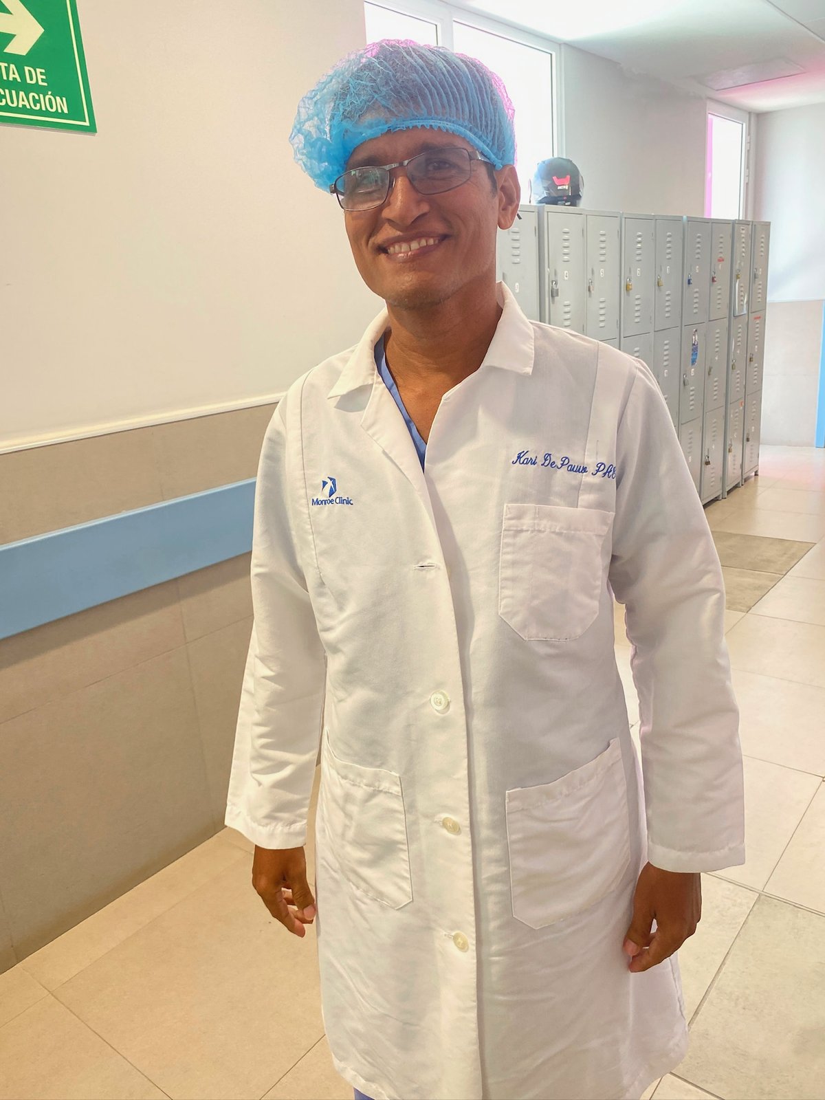 A clinician in a white lab coat and blue scrub cap smiling in a hospital hallway