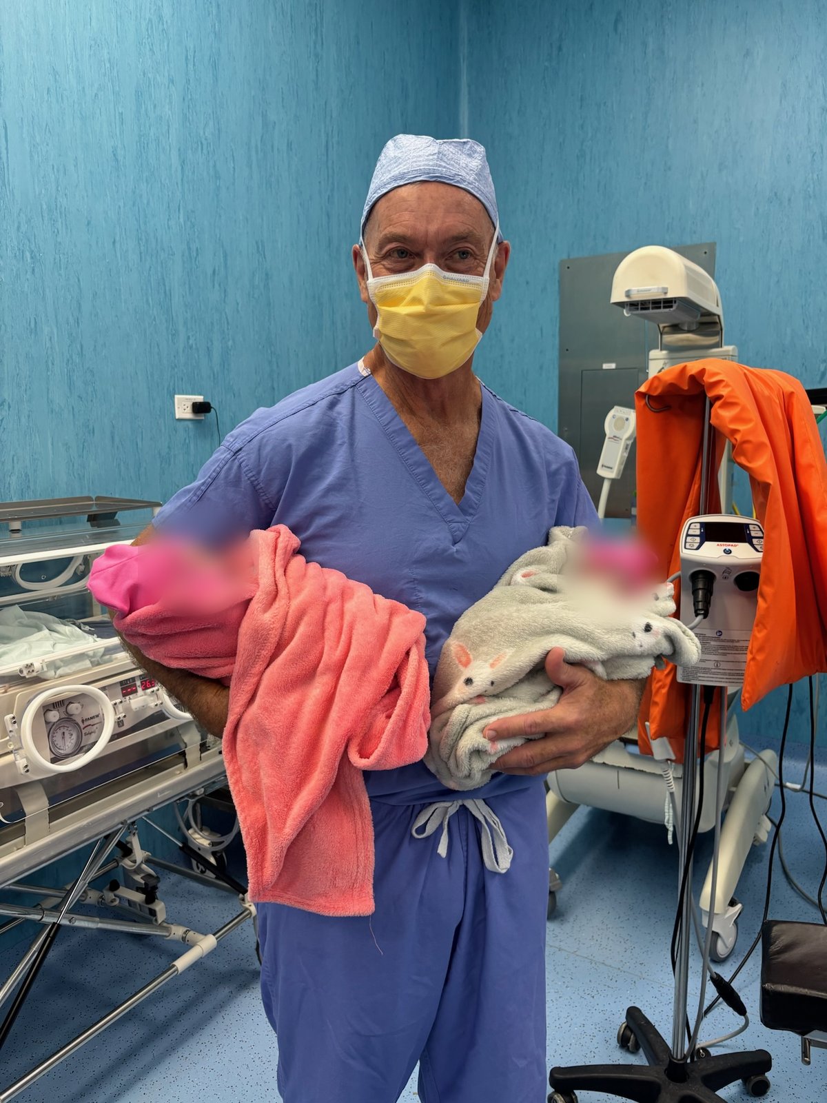 Thomas James Gallagher in blue scrubs and yellow mask holding two swaddled newborns
