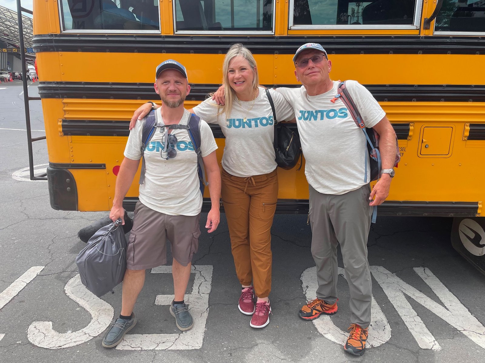 Three team members in matching JUNTOS shirts standing in front of a yellow school bus