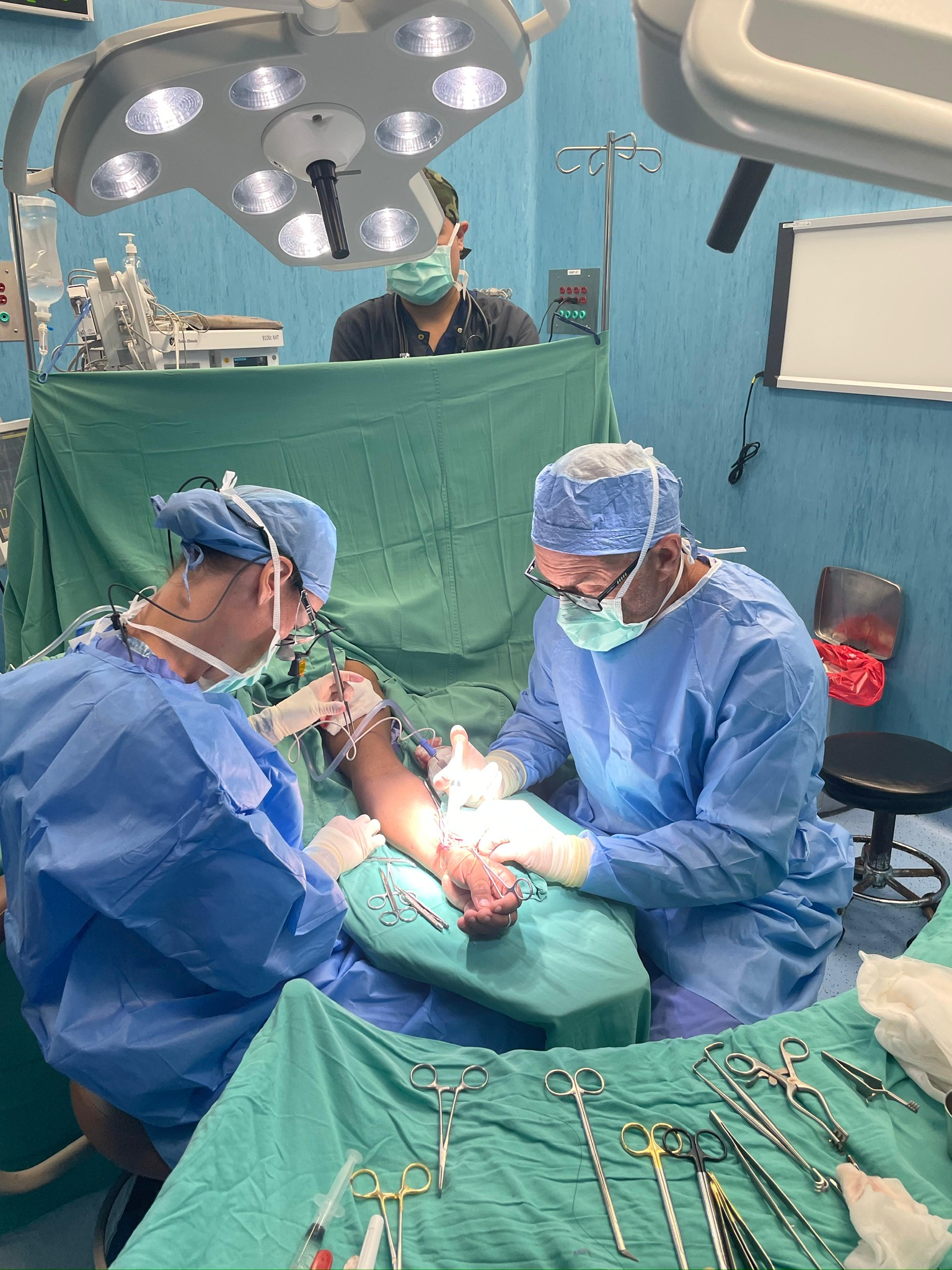 Surgical lights over two surgeons working on an arm, instruments laid out on green drapes