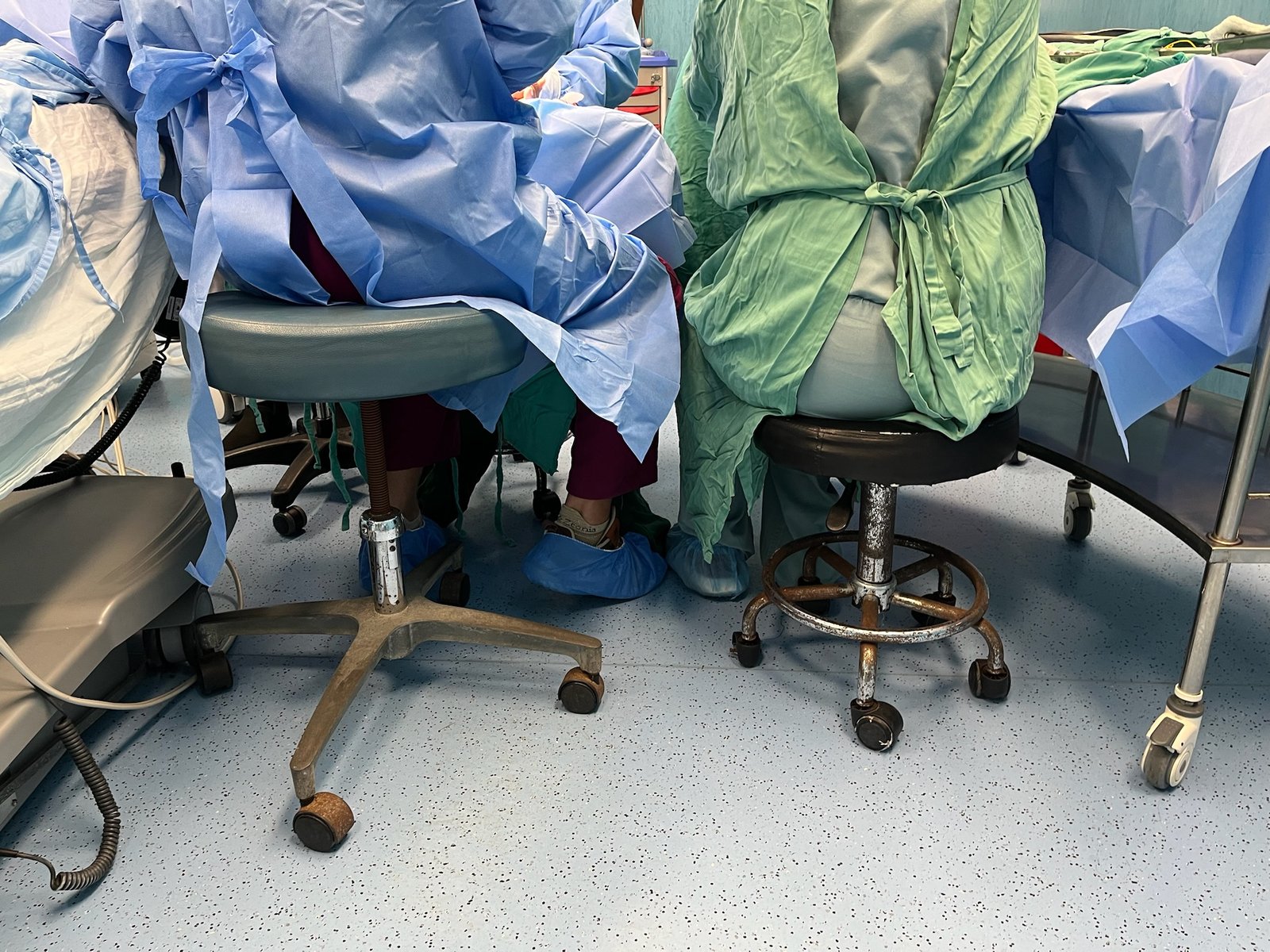 Two surgeons on rolling stools seen from behind in the operating room; no faces