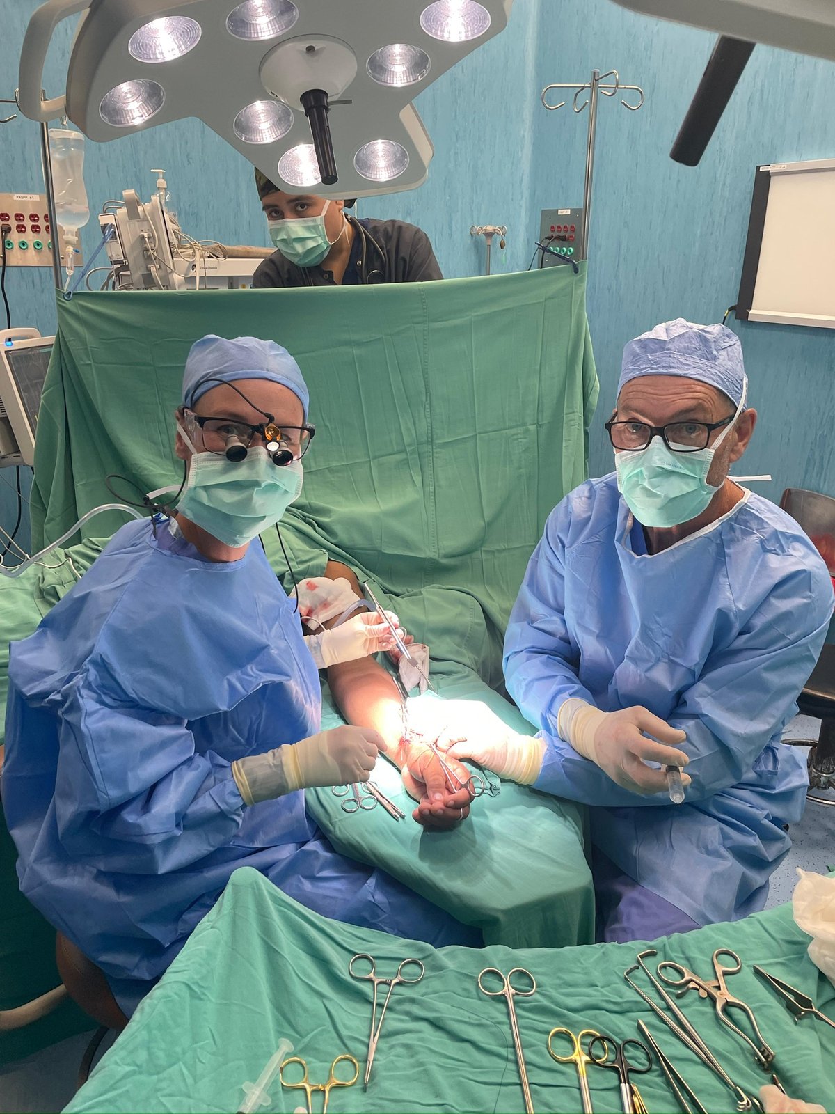 Two masked surgeons at the draped operating table looking up at the camera, instruments on green drapes in front; patient face not shown