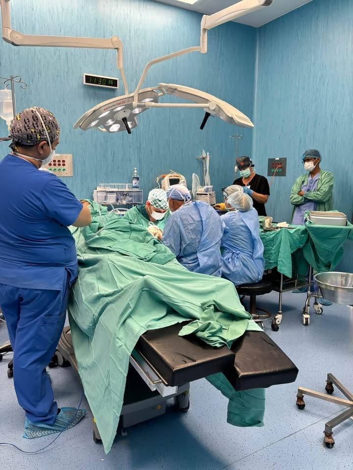 Surgical team gathered around a draped operating table under OR lights; patient face not shown