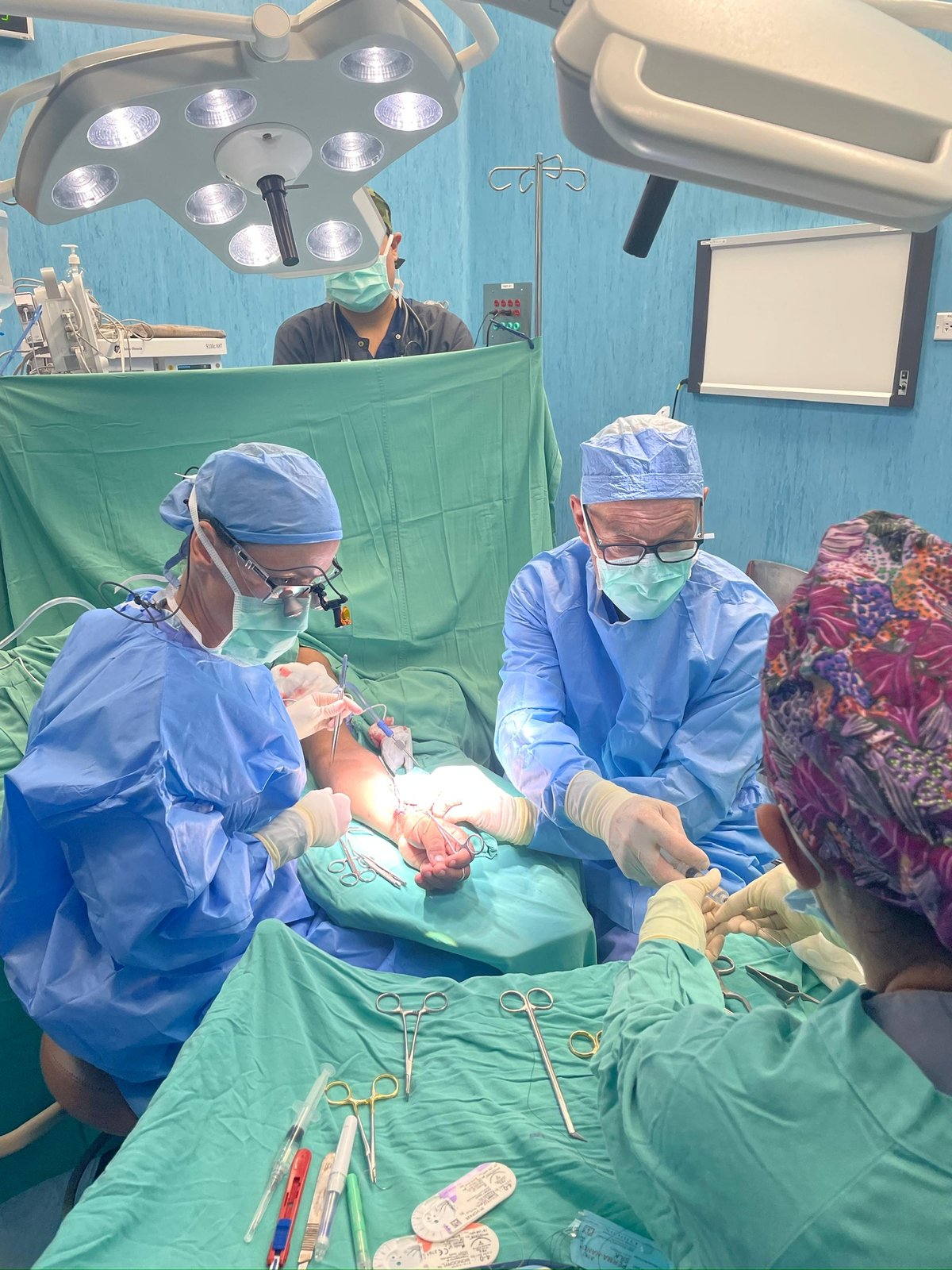 Under the OR lights: two surgeons at an arm, instrument tray in the foreground