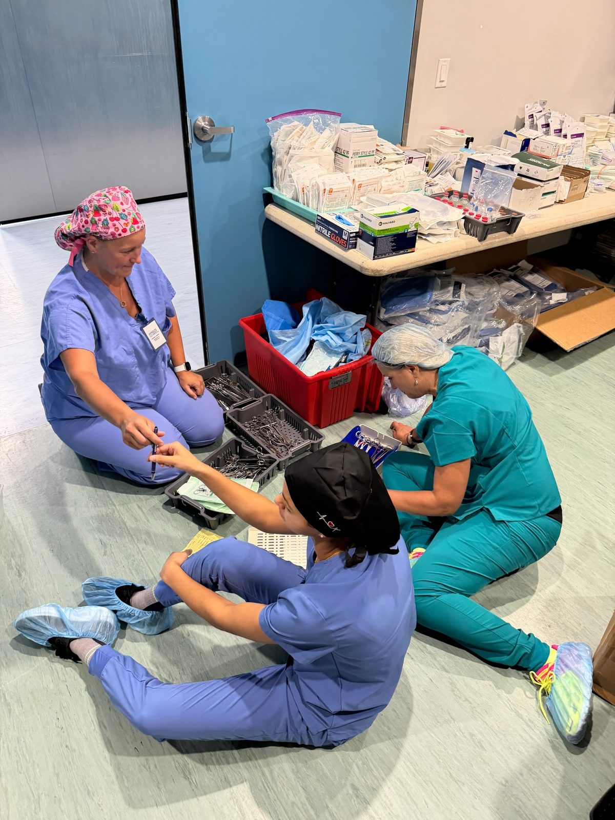 Three team members on the floor sorting instrument trays beside a red bin of drapes