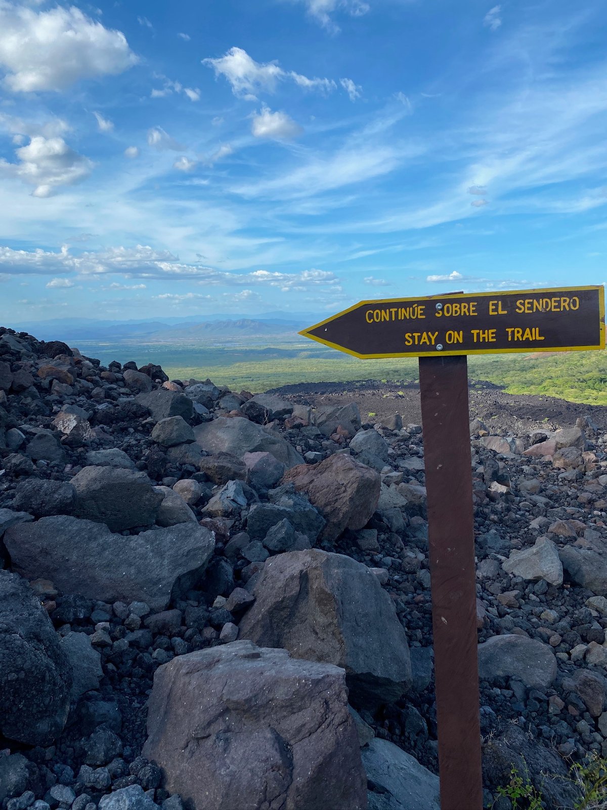 Bilingual stay-on-trail sign on volcanic rock with green valley and mountains beyond