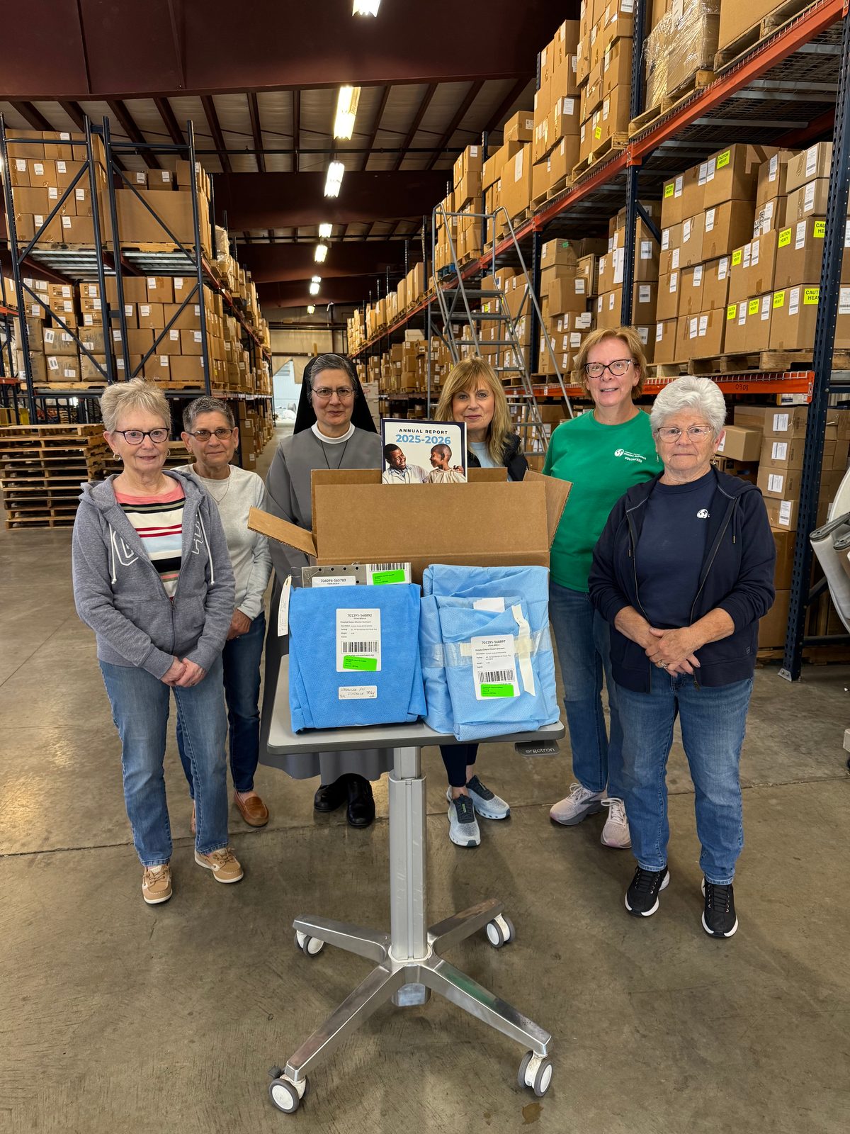 Six Hospital Sisters Mission Outreach volunteers in their warehouse, standing behind a cart with blue-wrapped donated surgical packs and a box of supplies